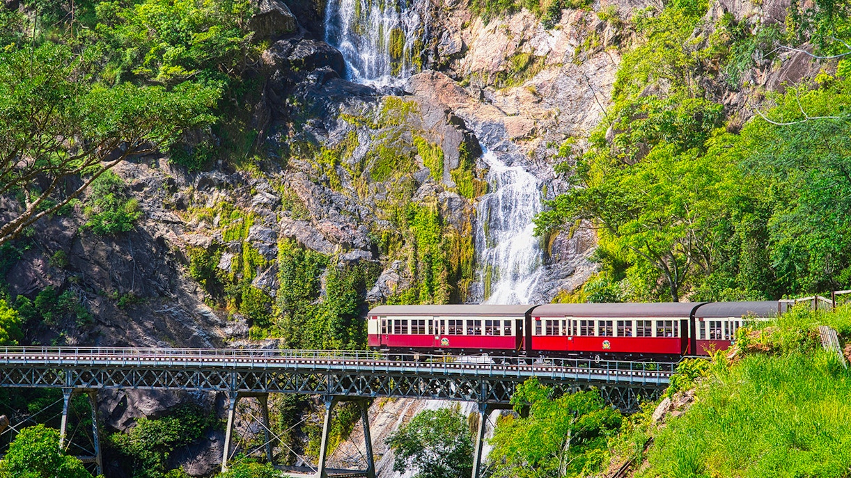 Kuranda Scenic Railway train crossing bridge with waterfall backdrop in Queensland, Australia.