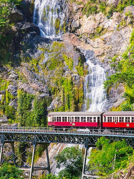 Kuranda Scenic Railway train crossing bridge with waterfall backdrop in Queensland, Australia.
