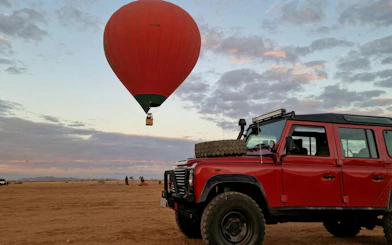 Hot air balloon ascending at sunrise near a red off-road vehicle in Marrakech desert.