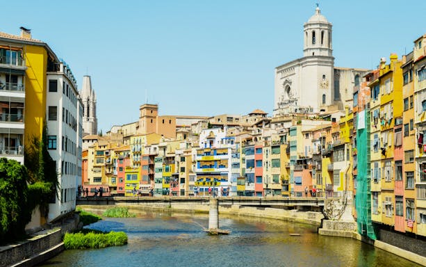 Colorful houses along the Onyar River in Girona, Spain with a cathedral in the background.