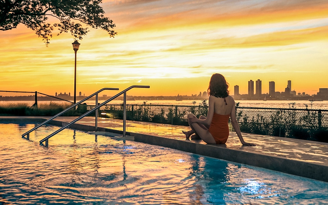 Person sitting by a pool at sunset with New York City skyline in the background.