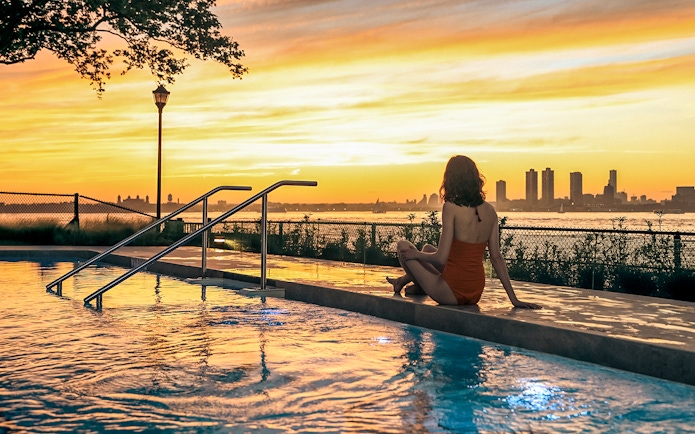 Person sitting by a pool at sunset with New York City skyline in the background.