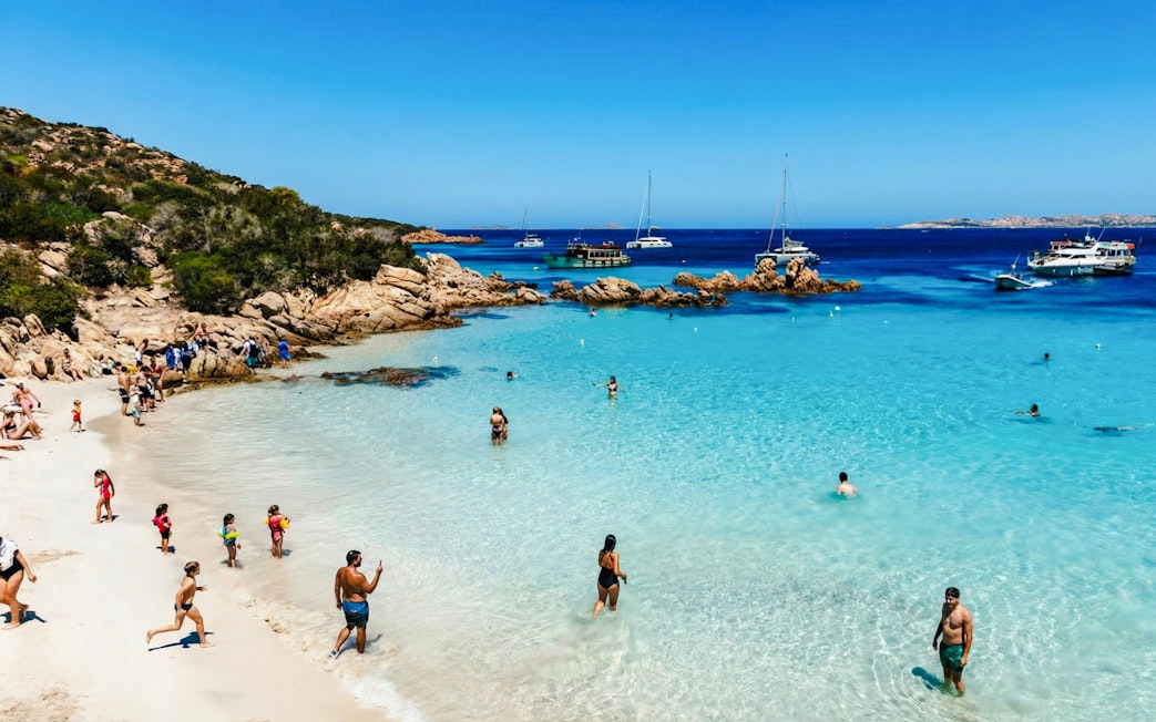 Motorboats anchored near a beach in La Maddalena Archipelago, people swimming and relaxing.