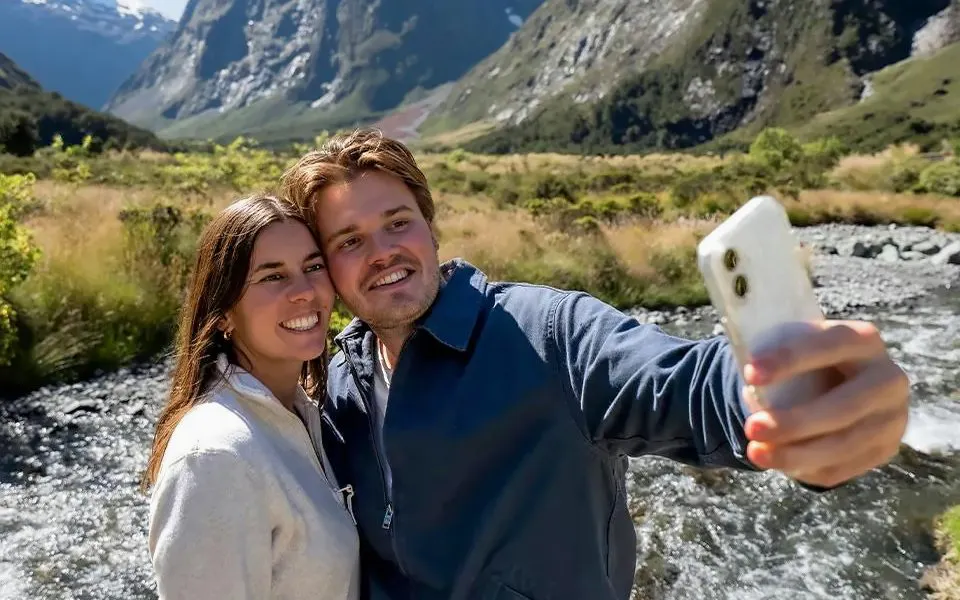 Couple taking a selfie by a river in Milford Sound, New Zealand, with mountains in the background.