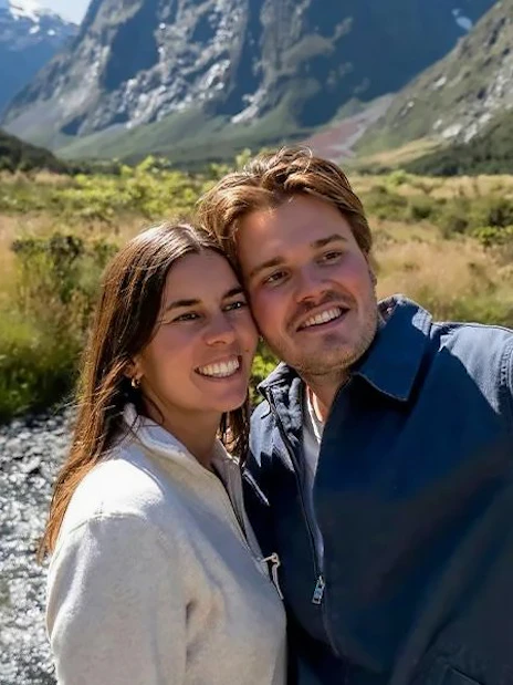 Couple taking a selfie by a river in Milford Sound, New Zealand, with mountains in the background.