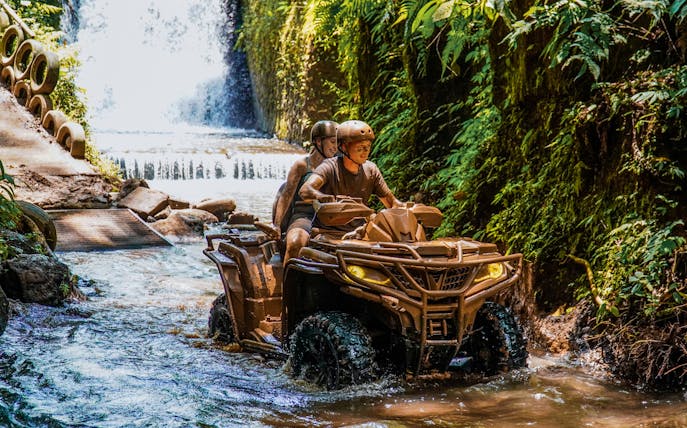 ATV riders navigating a stream near a waterfall in Ubud's lush jungle.