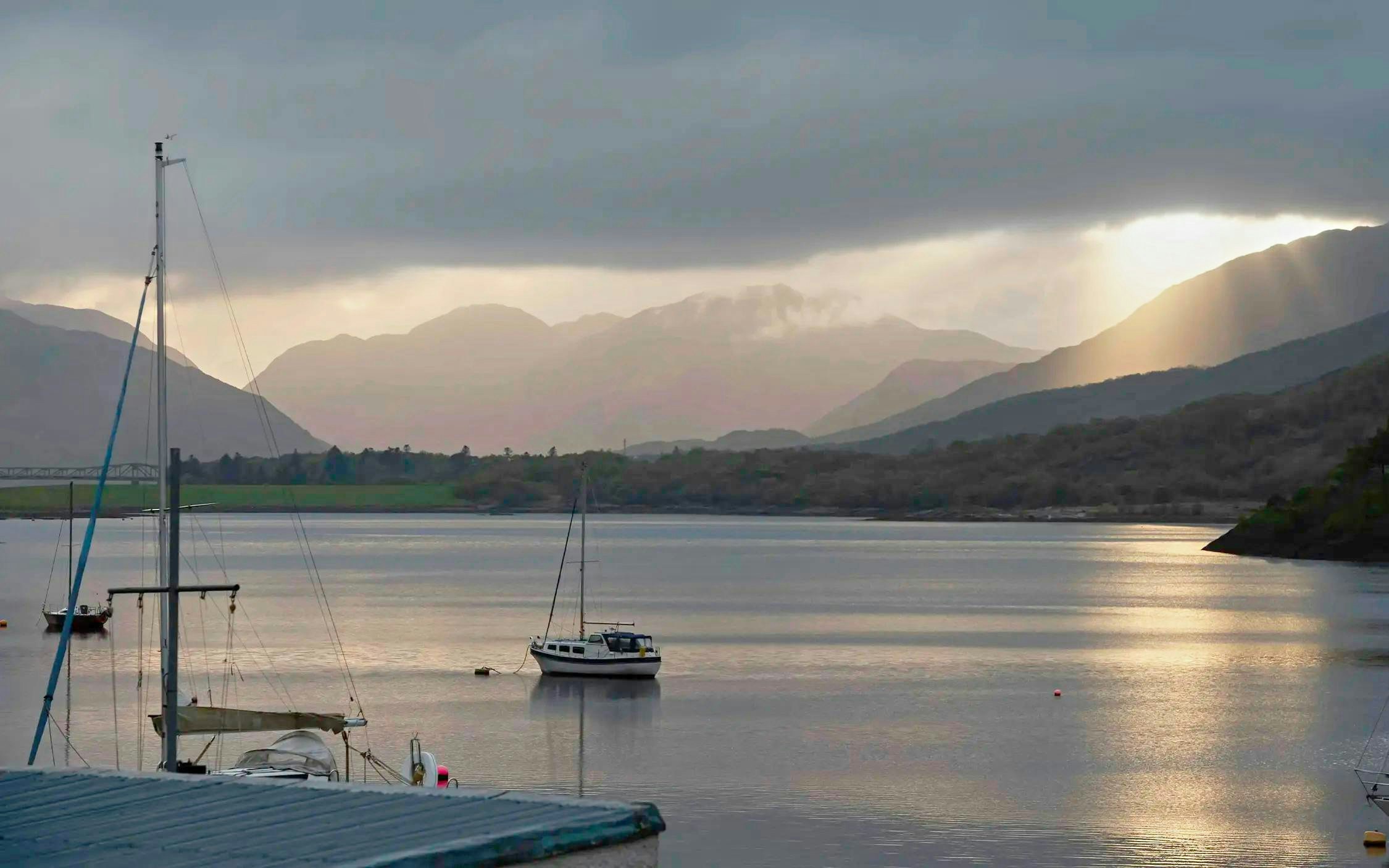 Sailboats on Loch Ness with sun rays breaking through clouds over surrounding hills.