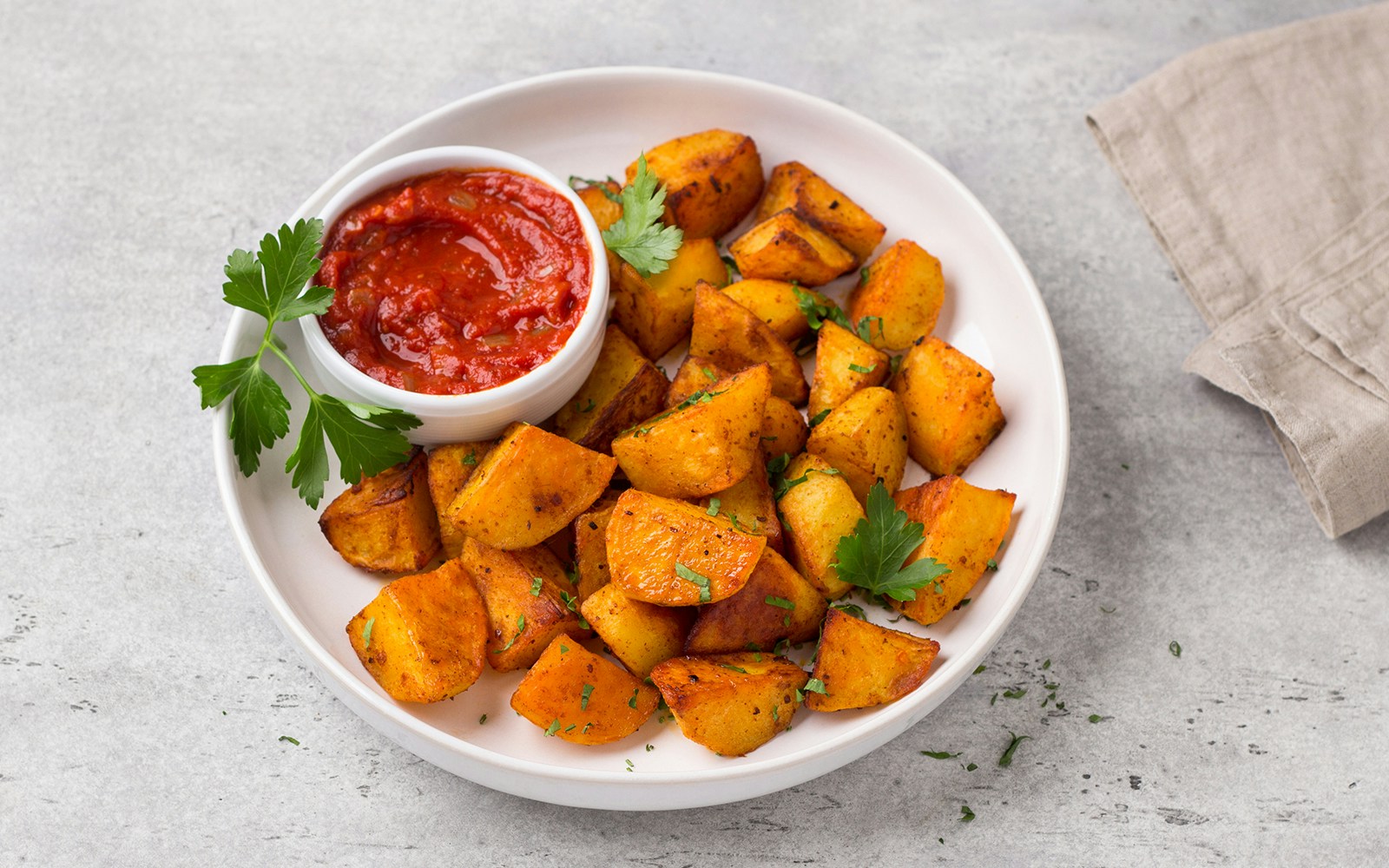 Traditional Spanish potato, patatas bravas with smoked paprika, spicy tomato sauce and parsley in a white bowl on gray stone