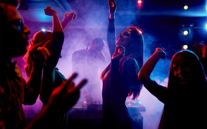 Women dancing energetically at a nightclub with a DJ in the background.
