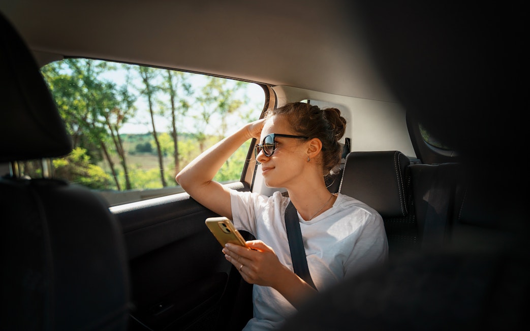 Young woman using smartphone in back seat of car, seatbelt fastened, looking out window.