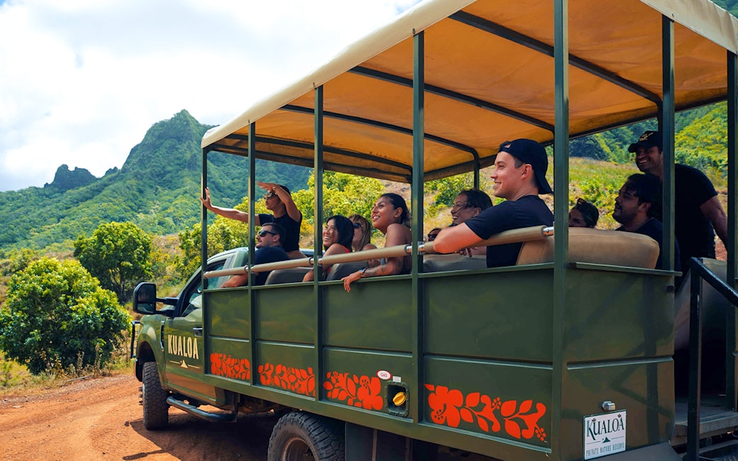 Tourists on a safari vehicle exploring Kualoa Ranch, Hawaii with lush mountains in the background.
