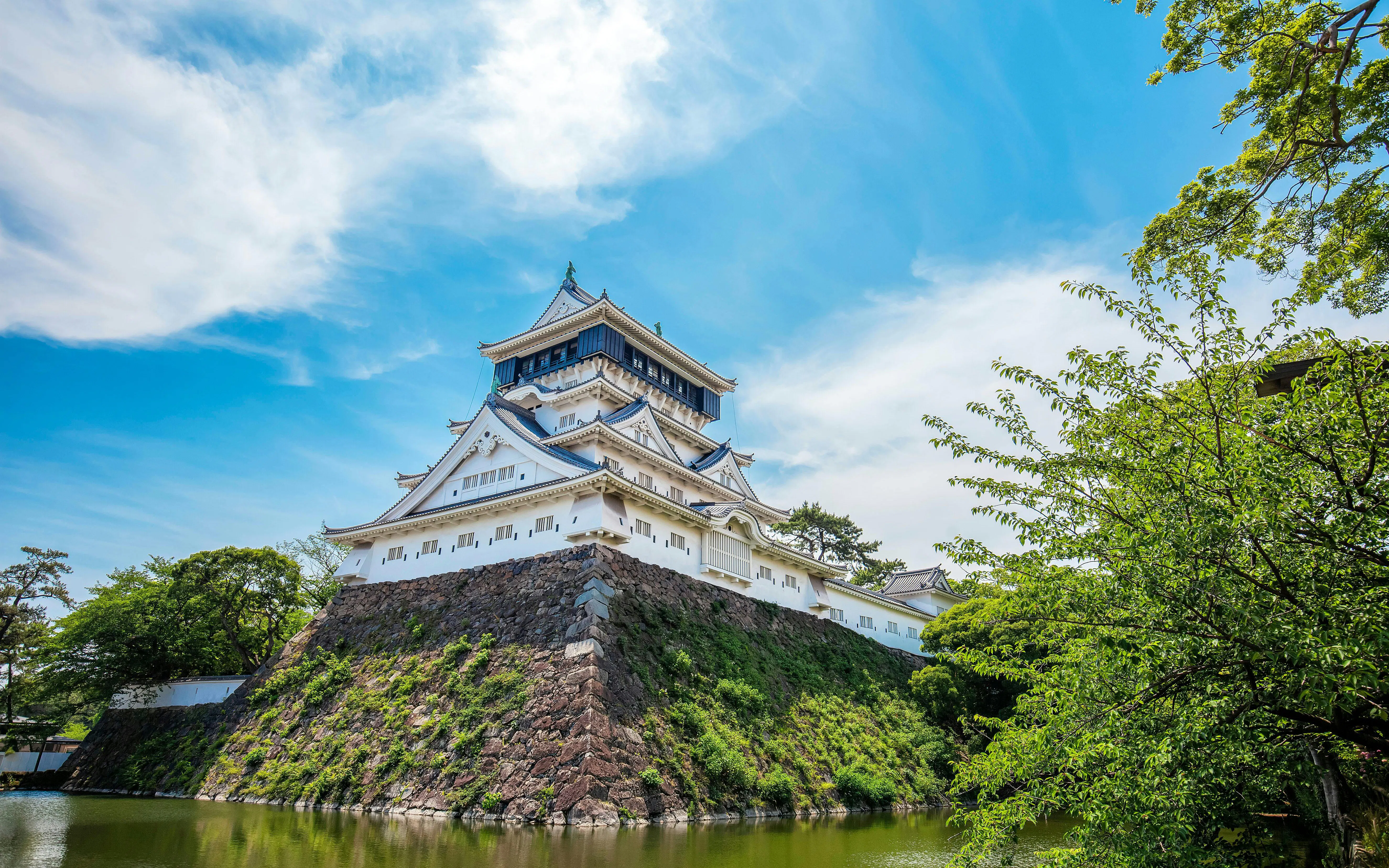 Kokura Castle in Japan with moat and trees under a blue sky.