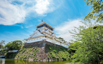 Kokura Castle in Japan with moat and trees under a blue sky.