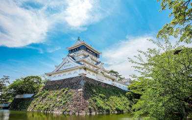 Kokura Castle in Japan with moat and trees under a blue sky.