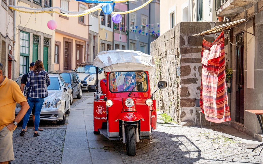 Red electric tuk-tuk on a narrow street in Porto, Portugal, with colorful buildings and decorations.