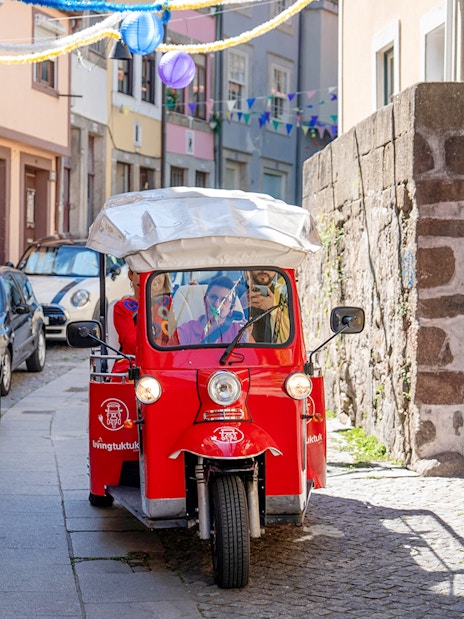 Red electric tuk-tuk on a narrow street in Porto, Portugal, with colorful buildings and decorations.