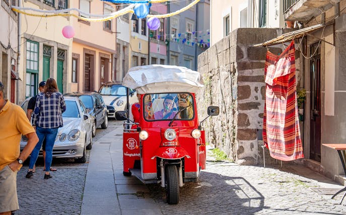 Red electric tuk-tuk on a narrow street in Porto, Portugal, with colorful buildings and decorations.