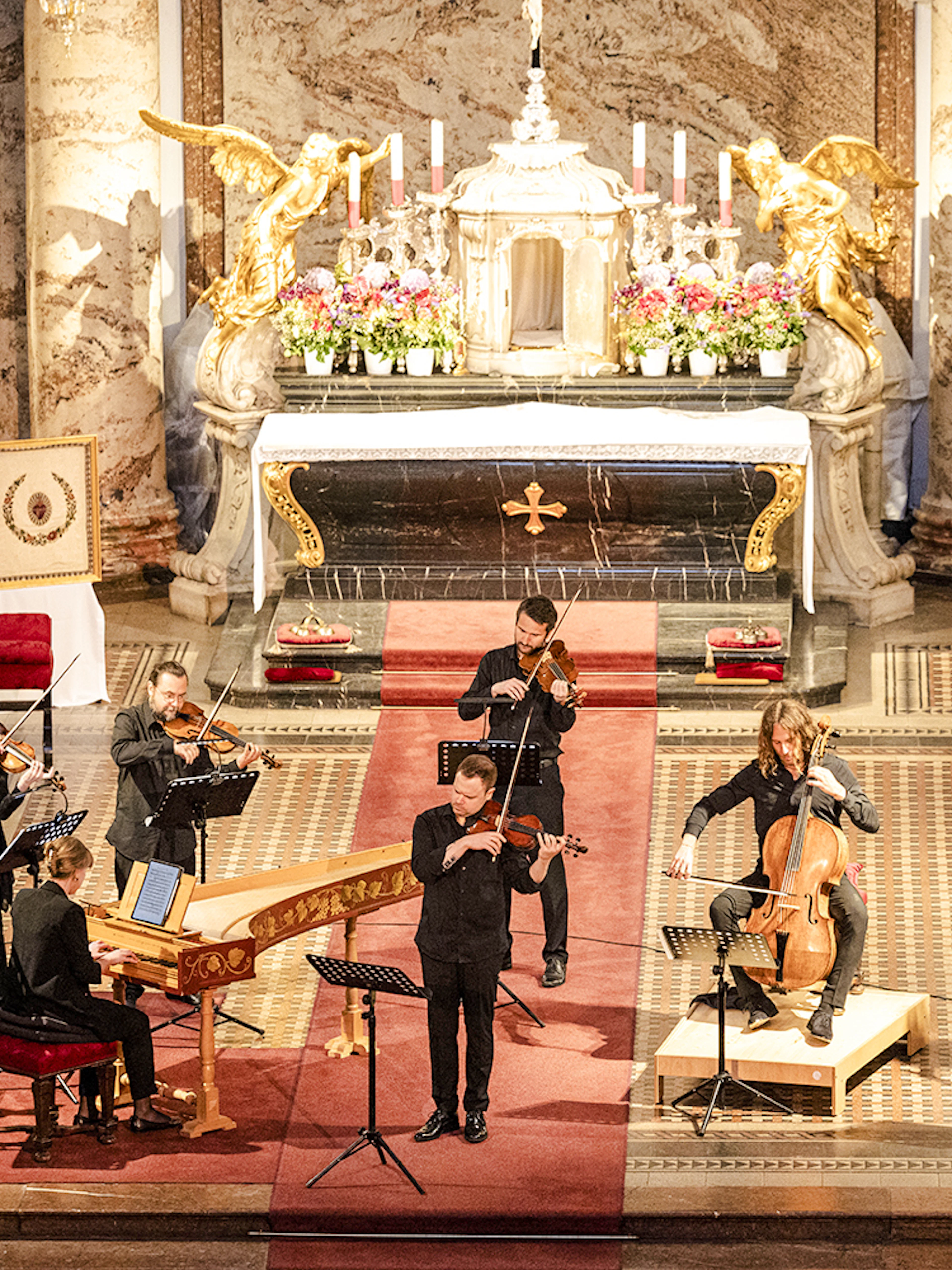 Musicians performing Vivaldi's Four Seasons inside a grand concert hall with ornate decor.