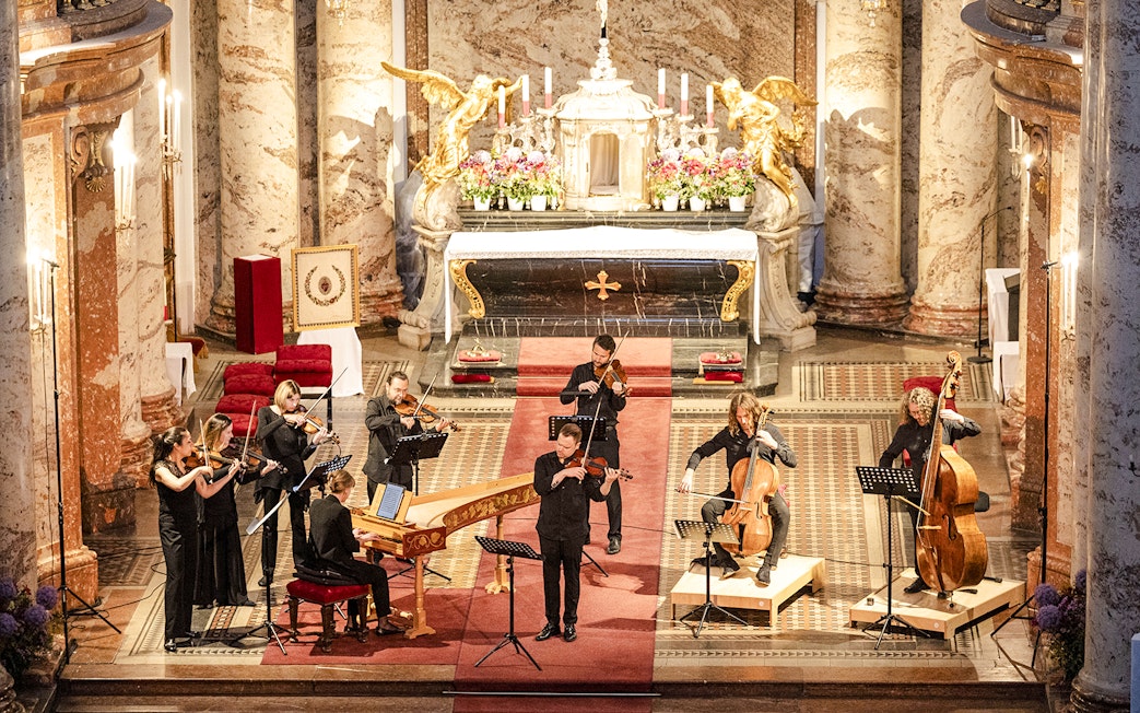Musicians performing Vivaldi's Four Seasons inside a grand concert hall with ornate decor.