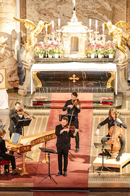 Musicians performing Vivaldi's Four Seasons inside a grand concert hall with ornate decor.