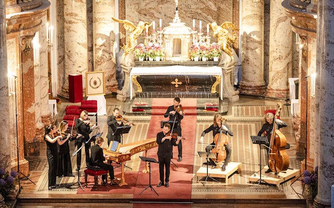 Musicians performing Vivaldi's Four Seasons inside a grand concert hall with ornate decor.