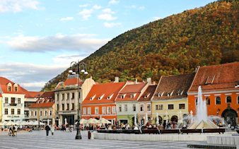 Brasov old town square with colorful buildings and fountain, framed by a forested hill.