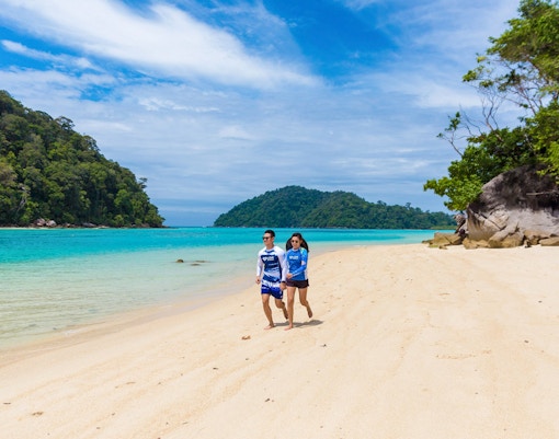 Couple walking on a sandy beach with turquoise water and lush greenery on Surin Island, Thailand.