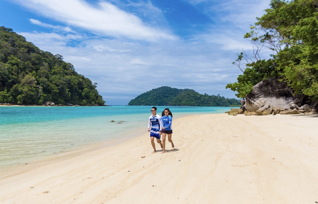 Couple walking on a sandy beach with turquoise water and lush greenery on Surin Island, Thailand.