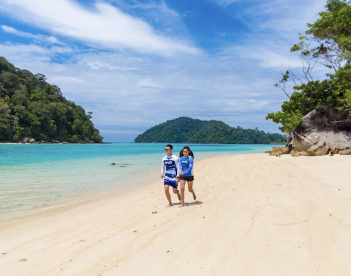 Couple walking on a sandy beach with turquoise water and lush greenery on Surin Island, Thailand.