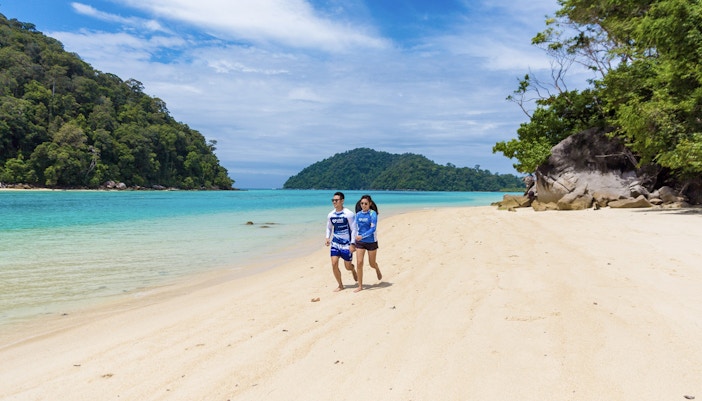 Couple walking on a sandy beach with turquoise water and lush greenery on Surin Island, Thailand.