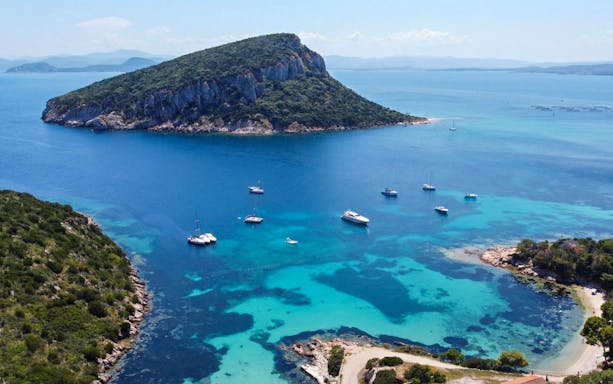 Boats anchored near Cala Moresca, Sardinia, with lush island and clear blue waters.