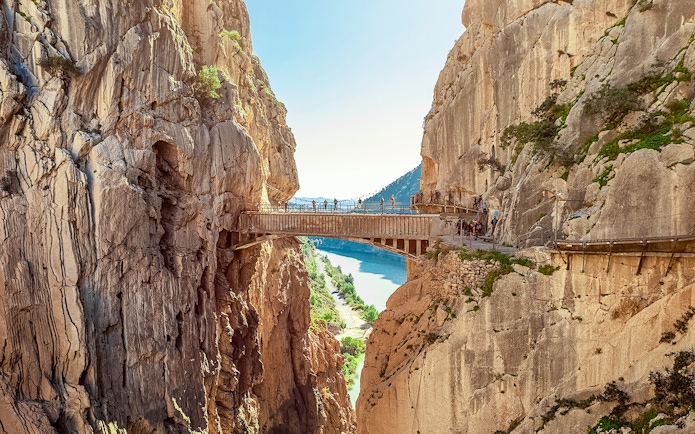 Hikers crossing a narrow bridge in Caminito del Rey, Spain, with cliffs and river below.