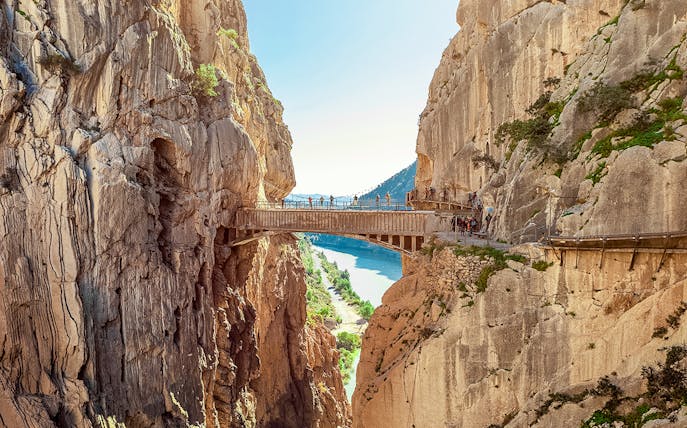 Hikers crossing a narrow bridge in Caminito del Rey, Spain, with cliffs and river below.