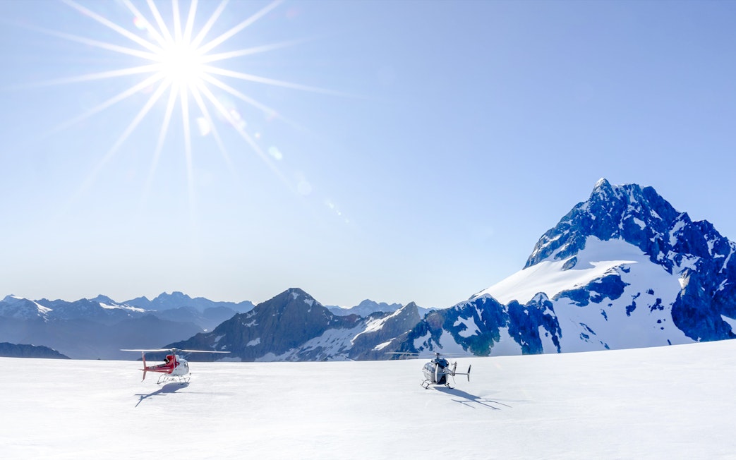 Two helicopters on a snow-covered glacier in the Southern Alps, Queenstown, under a bright sun.