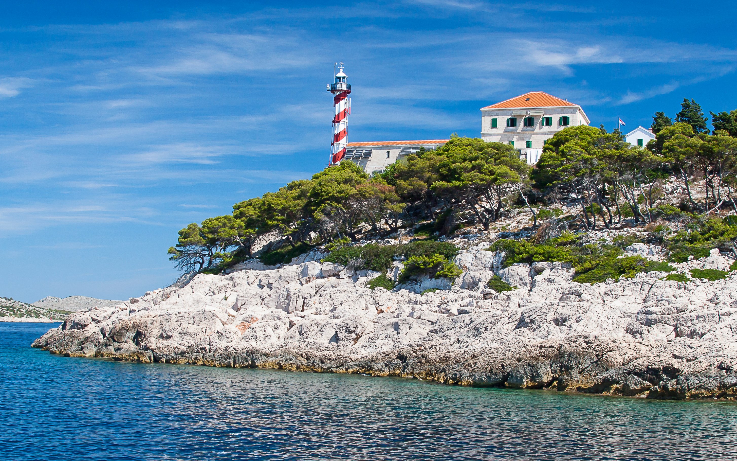 Lighthouse on rocky coast of National Park Kornati, Croatia, surrounded by trees and clear blue water.