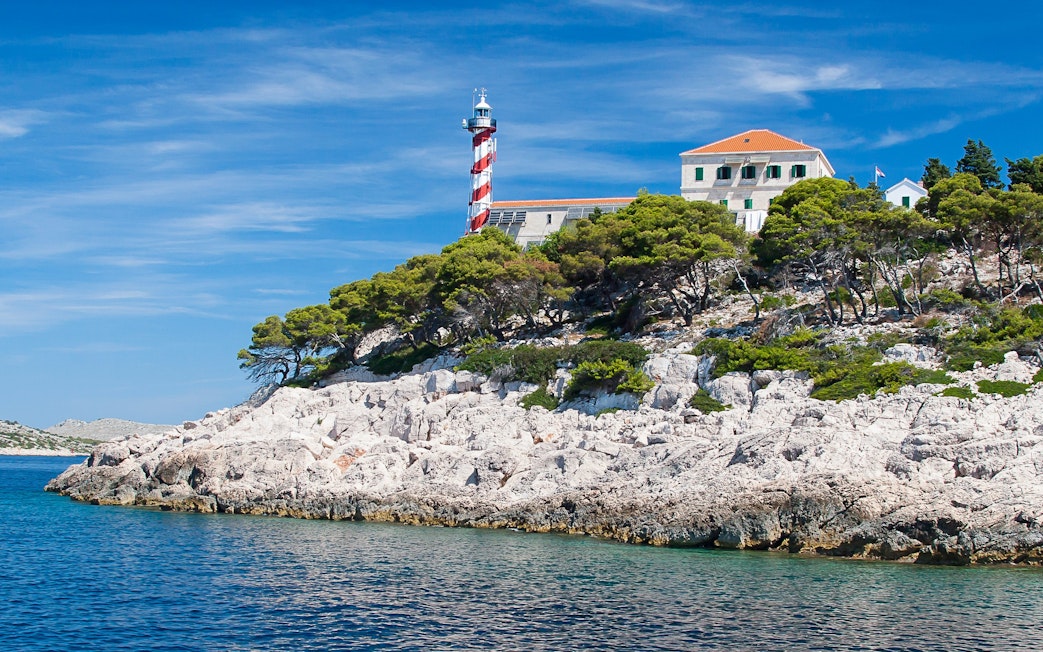 Lighthouse on rocky coast of National Park Kornati, Croatia, surrounded by trees and clear blue water.