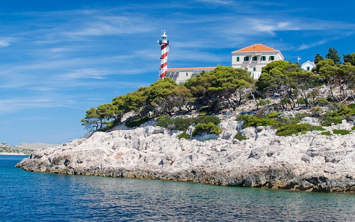 Lighthouse on rocky coast of National Park Kornati, Croatia, surrounded by trees and clear blue water.