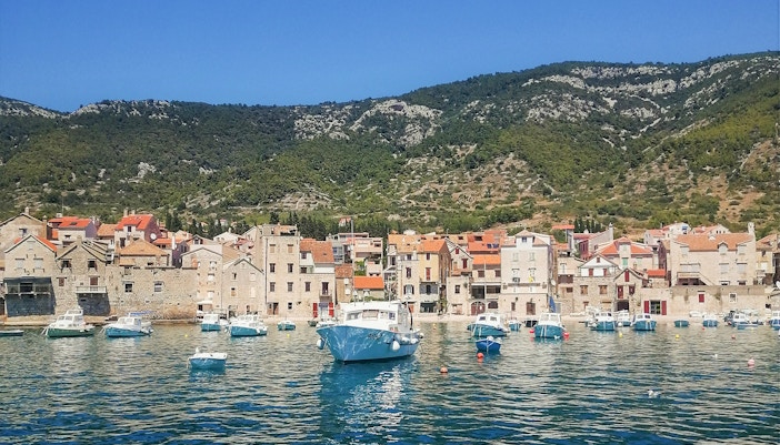 Boats in the harbor of Komiža, Vis Island with stone buildings and hills in the background.