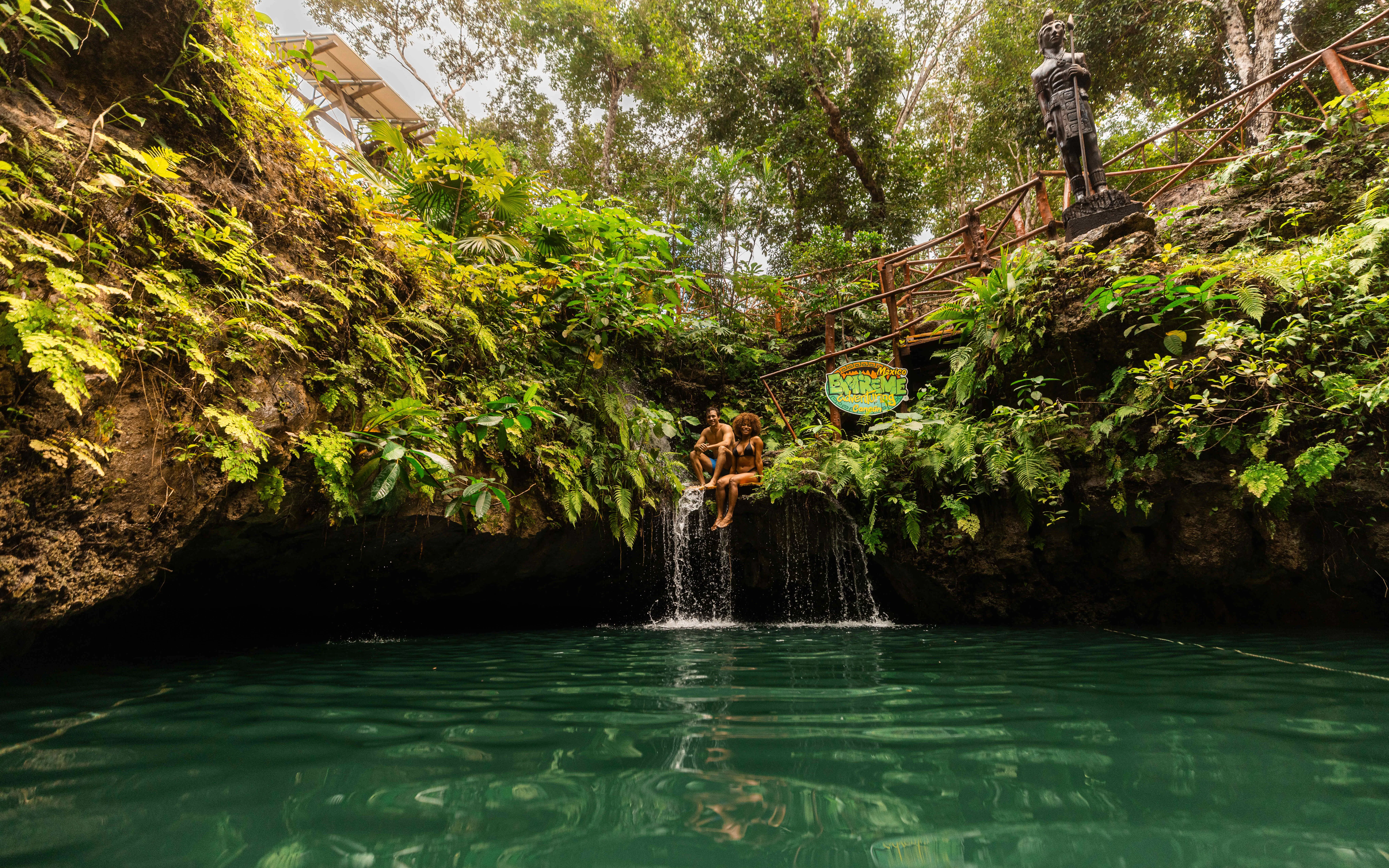 Tourists sitting by a waterfall in a lush cenote surrounded by greenery.
