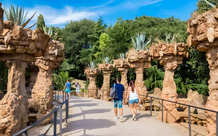 Visitors walking along a stone pathway in Park Güell, surrounded by unique rock columns and greenery.