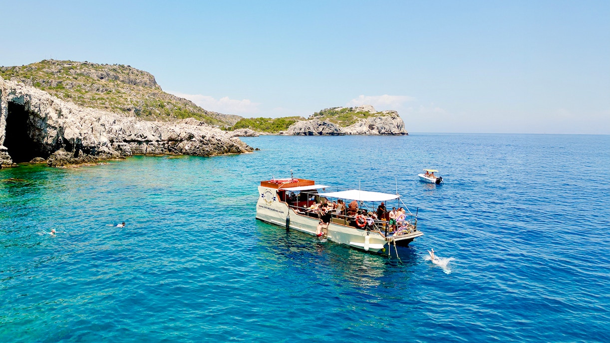 Boat tour near rocky caves in Rhodes with people swimming in the clear blue sea.