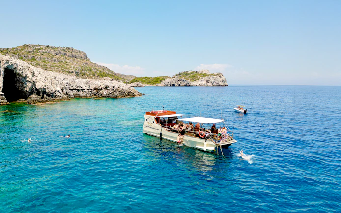 Boat tour near rocky caves in Rhodes with people swimming in the clear blue sea.