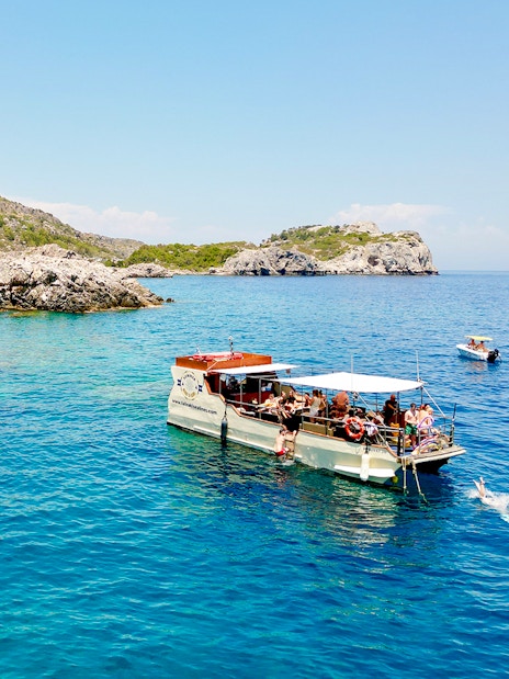 Boat tour near rocky caves in Rhodes with people swimming in the clear blue sea.
