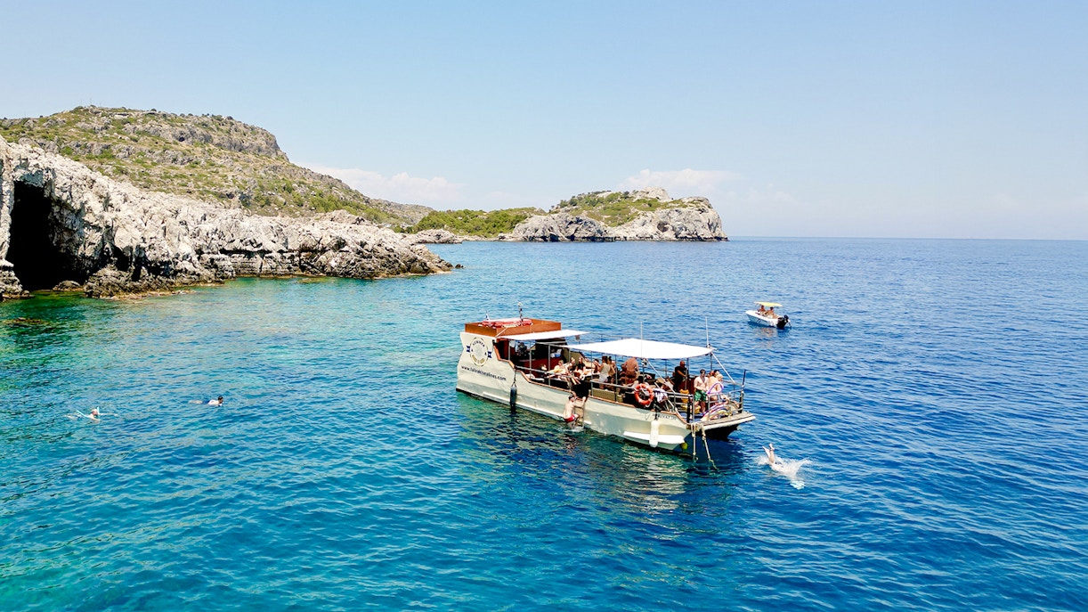 Boat tour near rocky caves in Rhodes with people swimming in the clear blue sea.