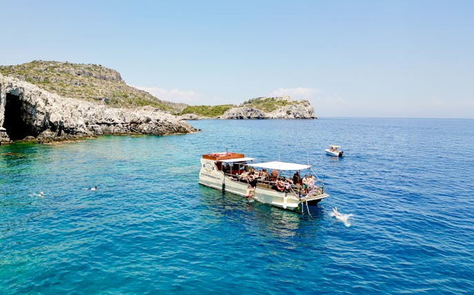 Boat tour near rocky caves in Rhodes with people swimming in the clear blue sea.