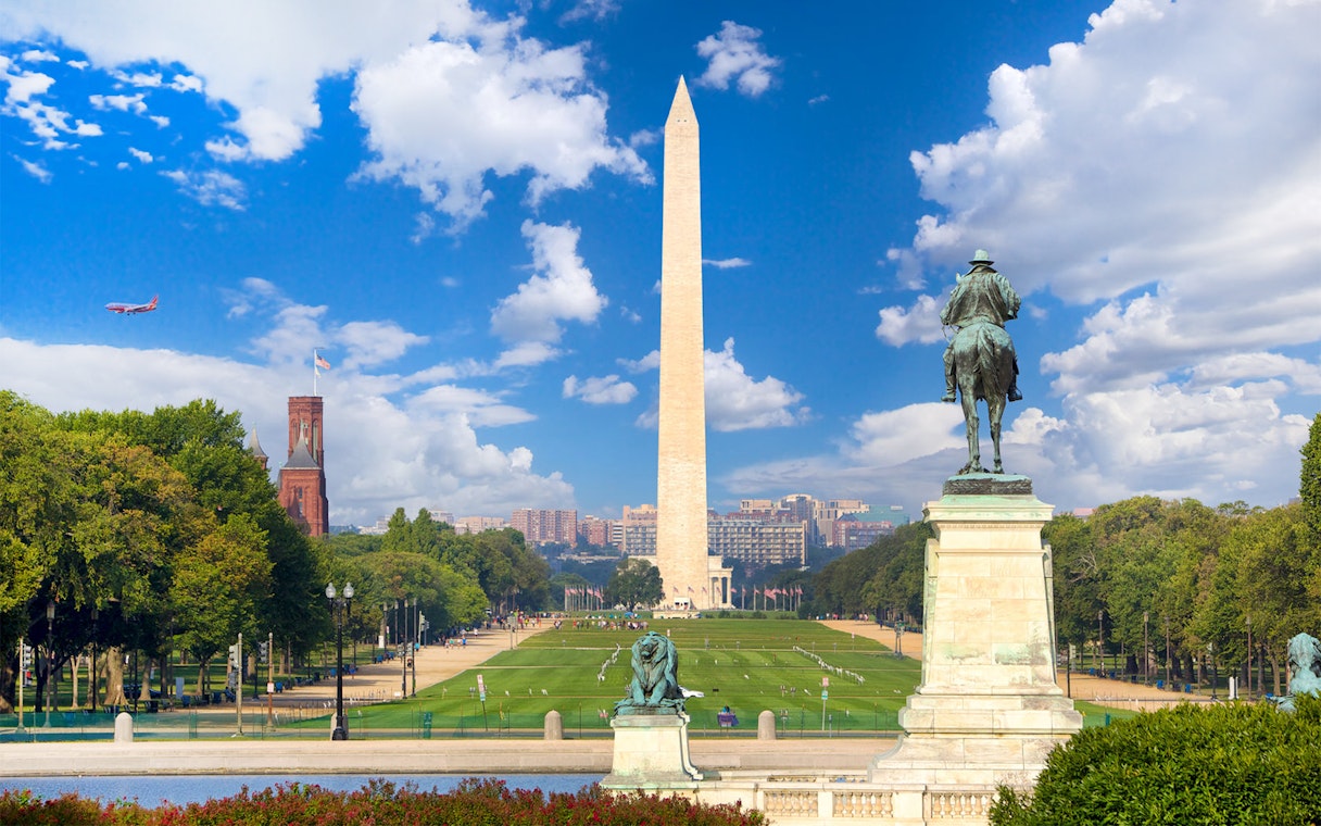 Washington Monument and statue at National Mall, Washington D.C., with clear sky and distant cityscape.
