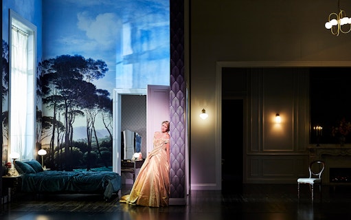 Performer in elegant gown on stage set of "La Traviata" with scenic backdrop.