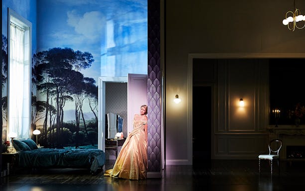 Performer in elegant gown on stage set of "La Traviata" with scenic backdrop.