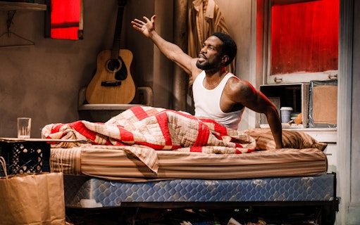 Man sitting on bed in a cluttered room with a guitar in the background, Topdog/Underdog theme.