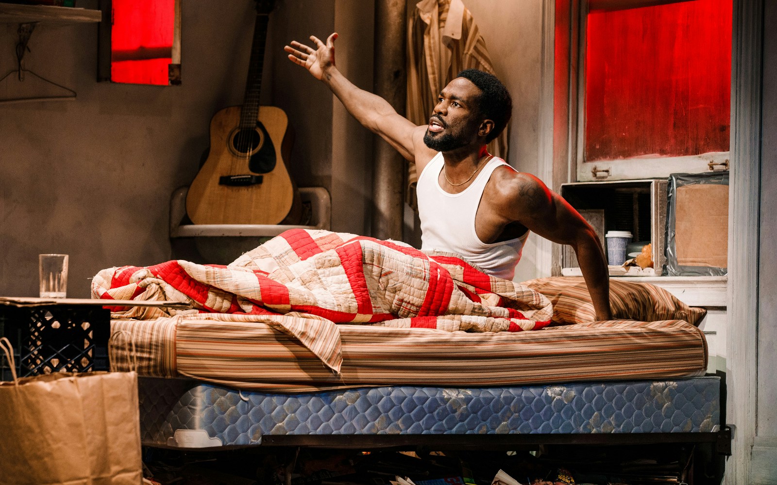 Man sitting on bed in a cluttered room with a guitar in the background, Topdog/Underdog theme.